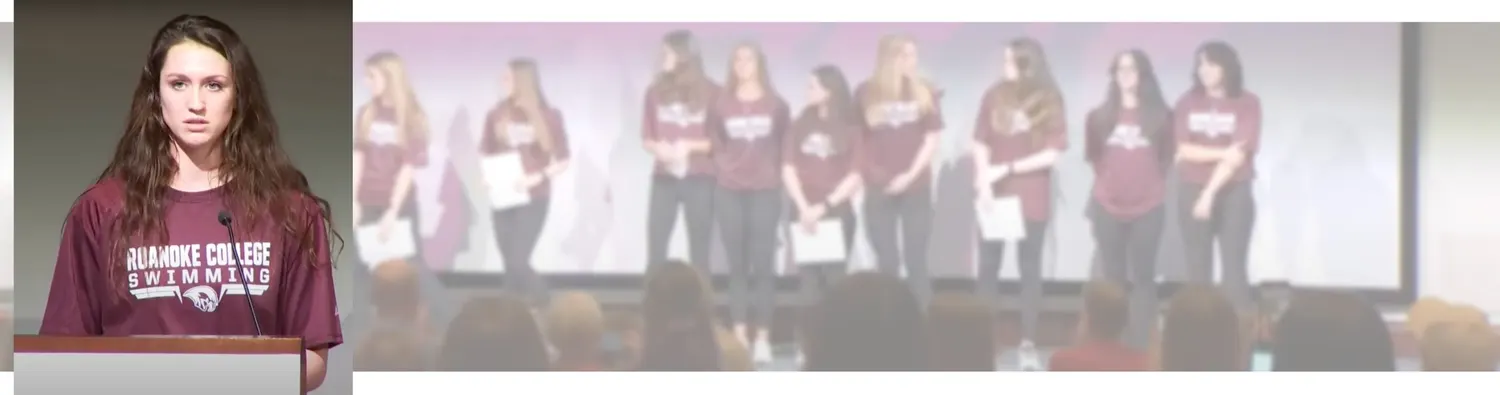 A Roanoke College swimmer speaks at a podium, with her teammates standing together behind her